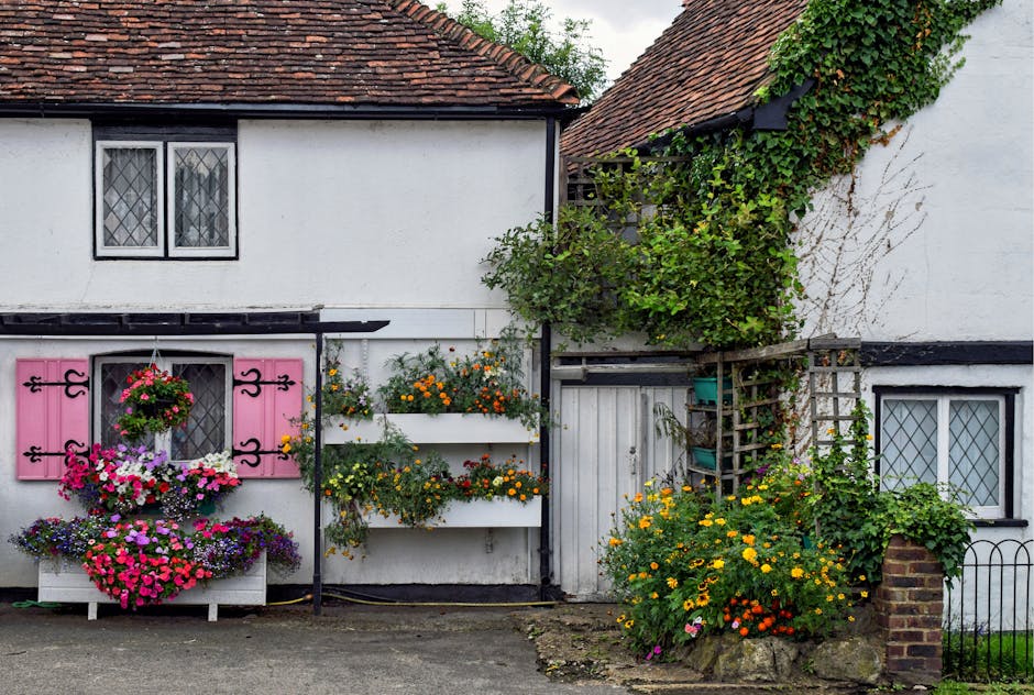 A charming white cottage with black trim and a tiled red roof, featuring vibrant flower displays in pink, purple, yellow, and orange. The window shutters are painted pink with black decorative hinges,