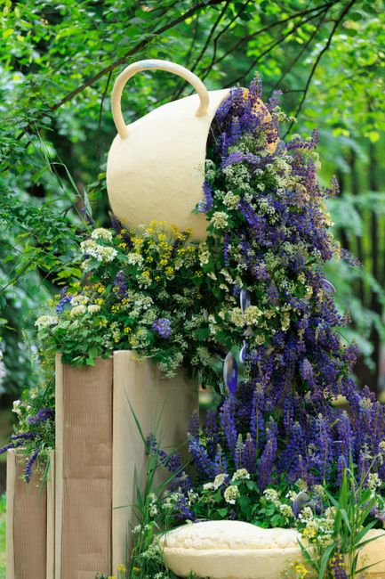 A floral display featuring a large, cream-colored, ceramic jug with a rounded handle set amidst lush greenery with vibrant green leaves and branches in the background. Surrounding the jug are abundant