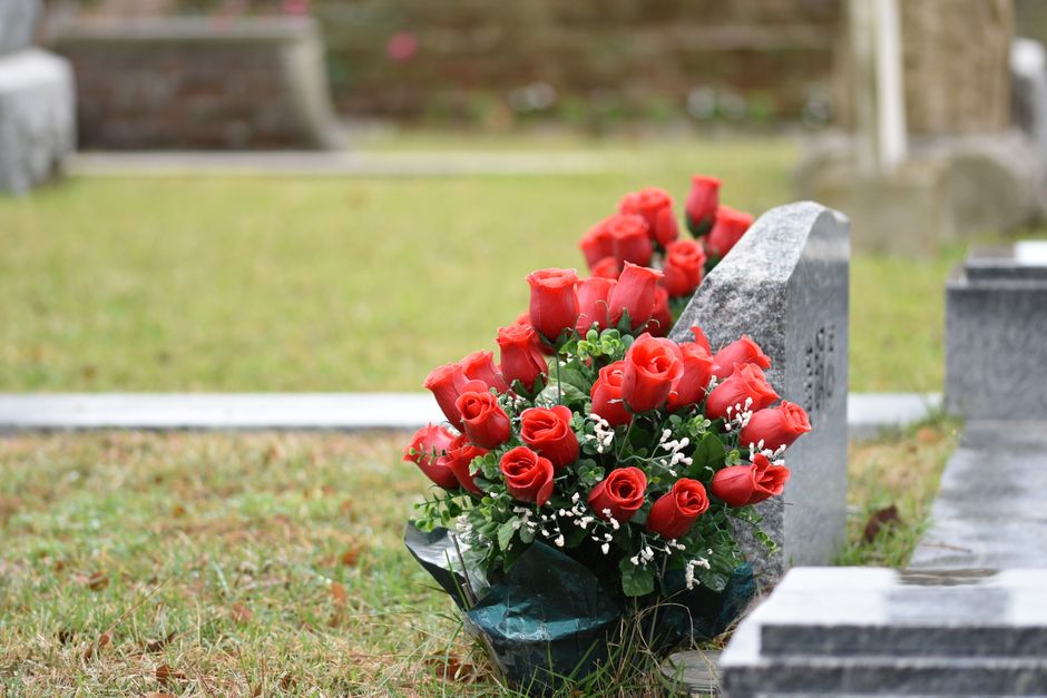 A bouquet of fresh red roses arranged with green foliage and small white filler flowers, wrapped in dark green floral paper. The bouquet is placed on the ground next to a grey gravestone in a cemetery