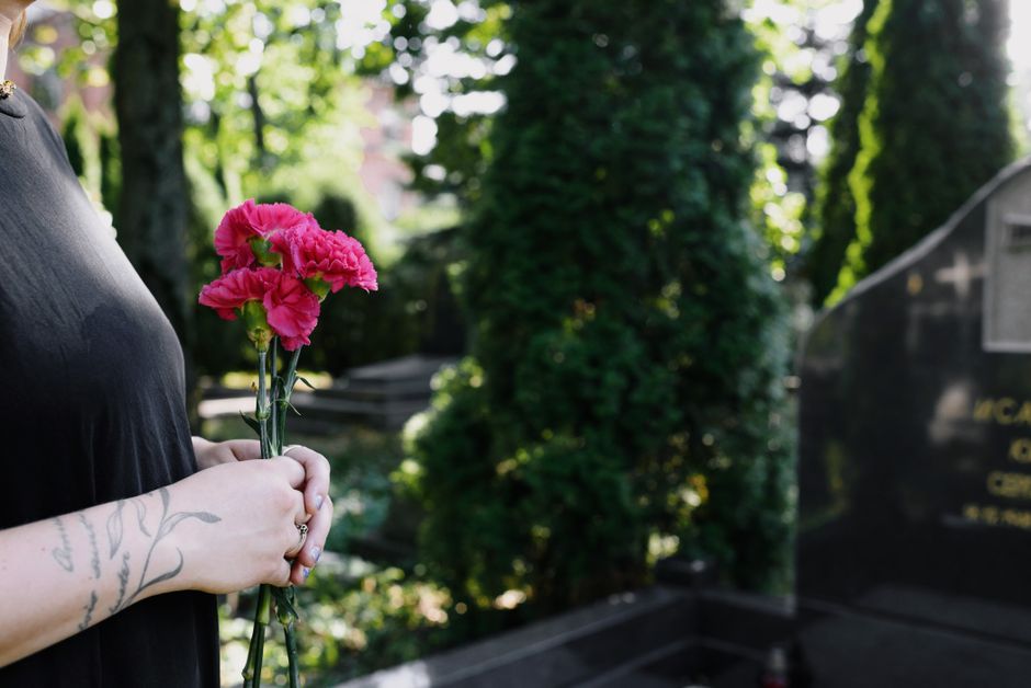 Close-up of a person holding a small bouquet of fresh pink carnations with ruffled petals and green stems, wrapped in clear plastic, at a memorial site. The individual has visible tattoos on their for