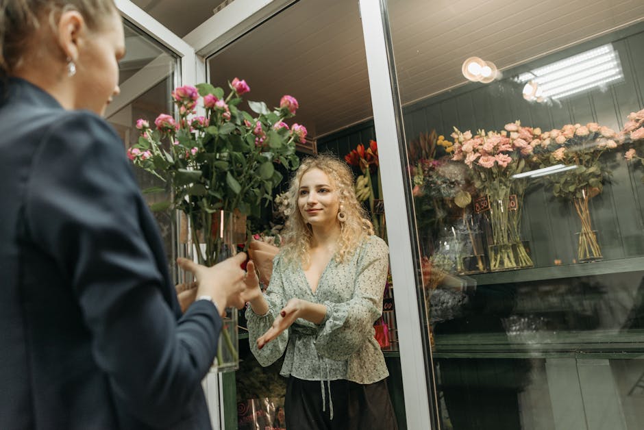 A floral arrangement scene at a flower shop, featuring a young woman with curly blonde hair, wearing a floral blouse and gesturing with her hands while receiving a bouquet of pink roses and greenery f