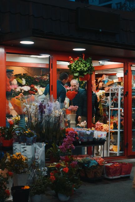 The image depicts the exterior of a flower shop on Wanstead High Street, illuminated by warm lighting, showcasing a vibrant display of fresh flowers arranged in baskets and vases on the pavement. Insi