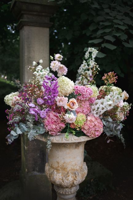 A large, lush floral arrangement in an antique stone urn, featuring a variety of fresh flowers including pale pink and white hydrangeas, soft peach roses, purple stocks, and delicate white and pink bl