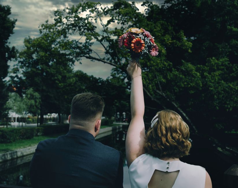 A woman with shoulder-length wavy red hair, wearing a white dress with a small open back, is holding a vibrant bouquet of mixed flowers high above her head with her right hand. The bouquet includes or
